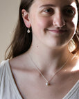 A close-up lifestyle shot of a woman wearing a delicate silver chain necklace with a single white pearl pendant, along with matching stud earrings.