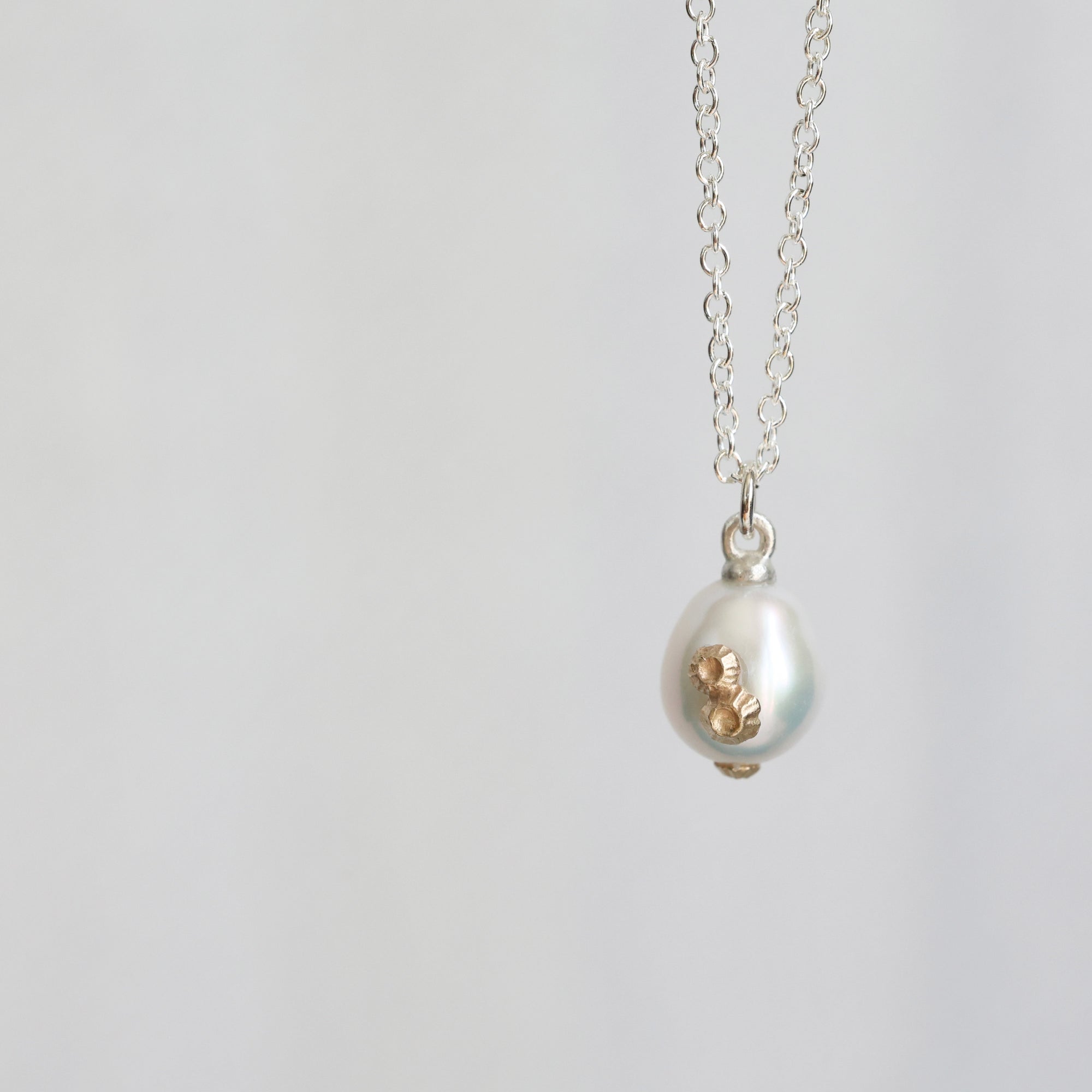 A silver chain necklace with a teardrop-shaped white pearl pendant featuring a textured gold accent on the side, shown against a plain light gray background.