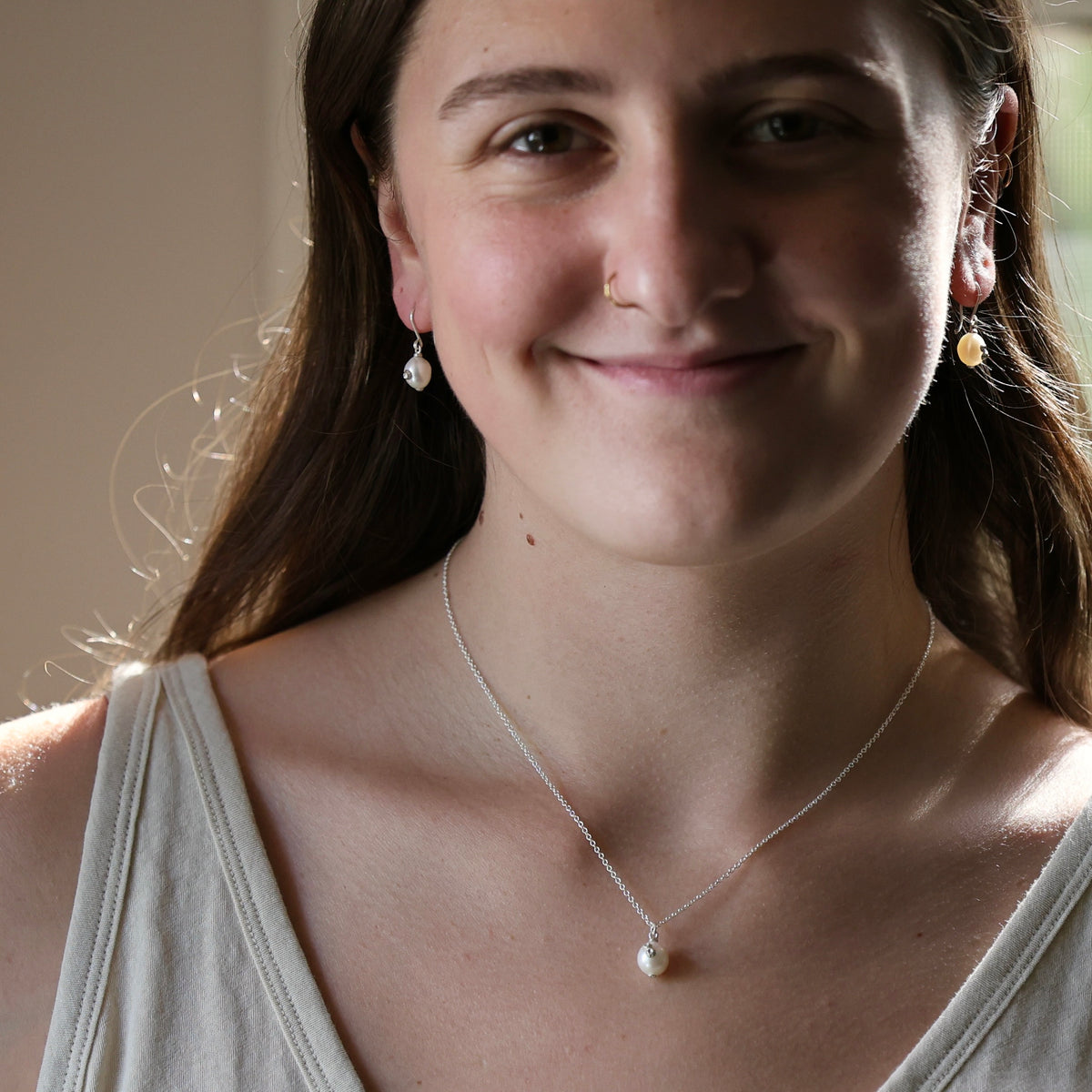 A smiling young woman with long brown hair wearing a delicate silver chain necklace with a small white pearl pendant and matching drop earrings, shown in a close-up portrait.