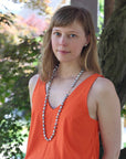 Woman wearing an orange sleeveless top with a long necklace, standing outdoors with trees in the background.