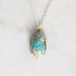 A silver chain necklace with an oval turquoise pendant set in a gold-toned prong setting, resting on a light marble surface.