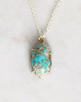 A silver chain necklace with an oval turquoise pendant set in a gold-toned prong setting, resting on a light marble surface.