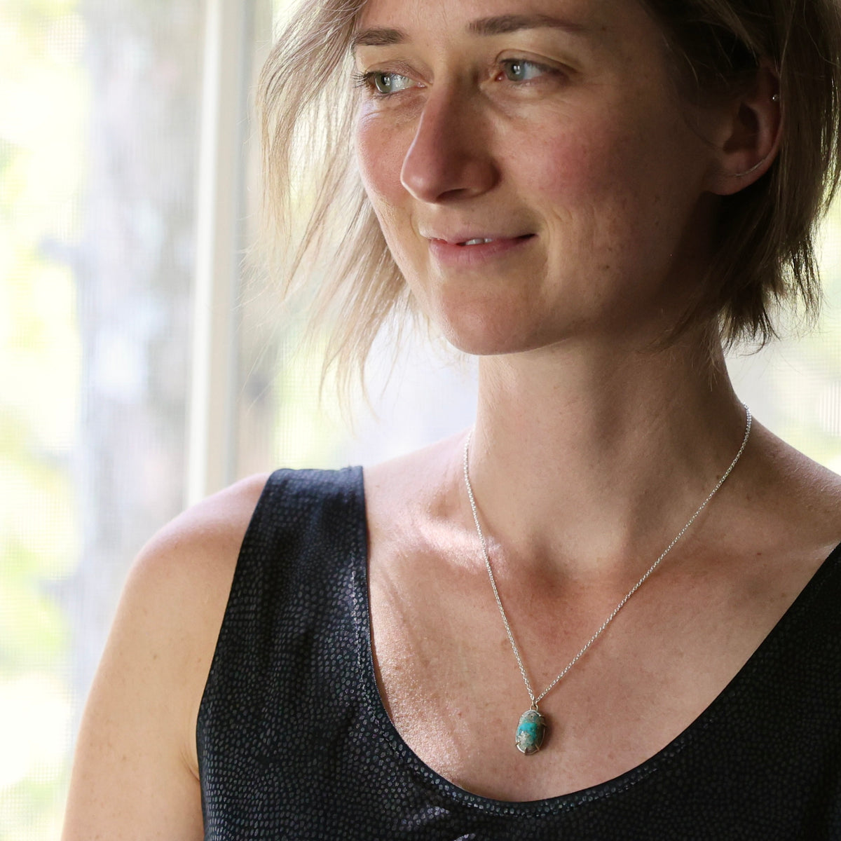 A woman wearing a delicate silver chain necklace with a small, oval turquoise-colored stone pendant, photographed in soft, natural light near a window.