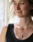 A woman wearing a delicate silver chain necklace with a small, oval turquoise-colored stone pendant, photographed in soft, natural light near a window.