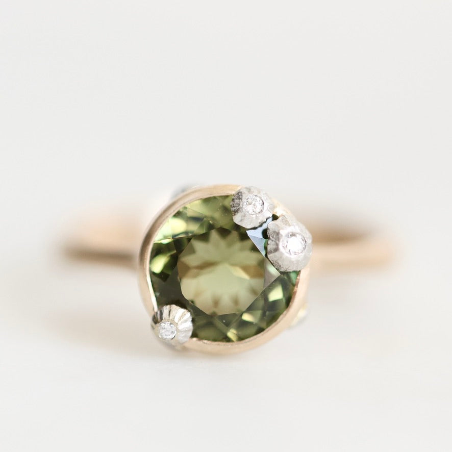 A close-up shot of a ring featuring a large oval green gemstone in a gold setting, accented by three small round clear stones on the upper right, against a plain light background.