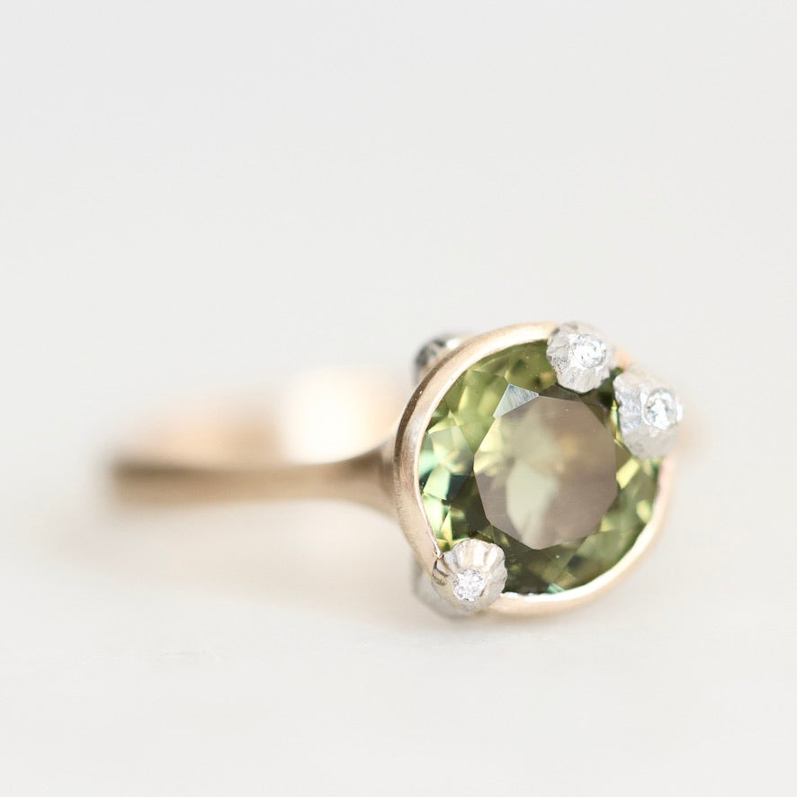 A gold ring featuring a large oval green gemstone in a prong setting, accented with three small round clear stones, shown on a light gray background.