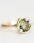 A gold ring featuring a large oval green gemstone in a prong setting, accented with three small round clear stones, shown on a light gray background.