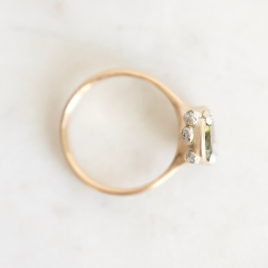 A gold ring with a small cluster of clear gemstones on the setting, photographed from above on a light off-white marble surface.