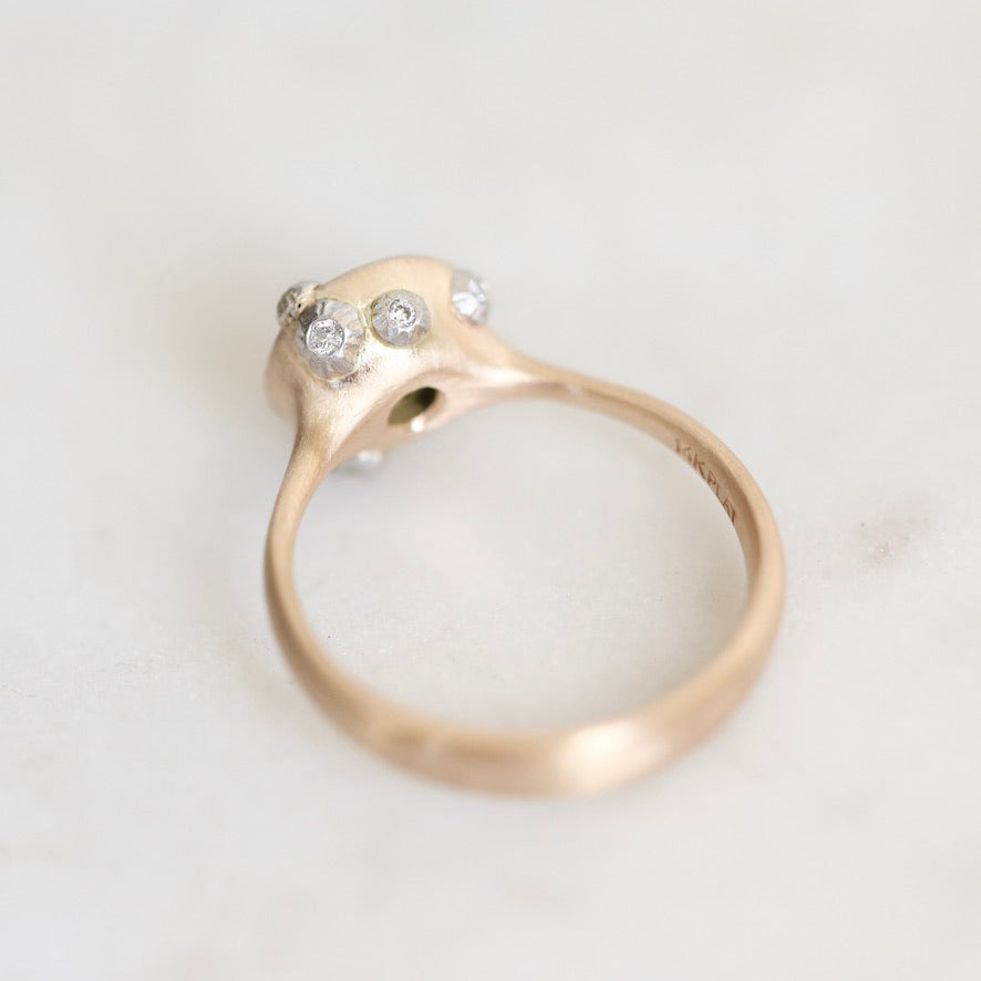 A close-up shot of a gold ring featuring a unique organic-shaped setting with three small clear gemstones, photographed on a light, neutral background.