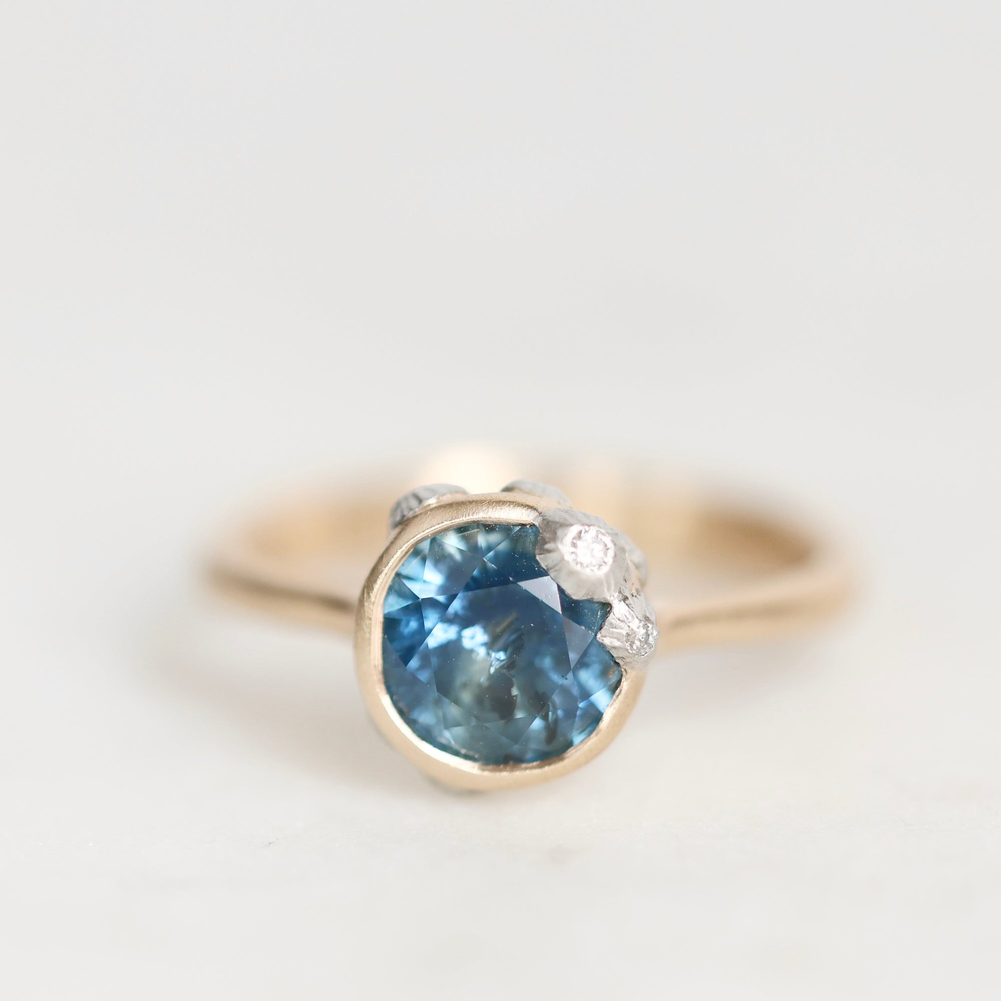A gold ring featuring a round-cut blue sapphire center stone and small clear accent diamonds, photographed on a clean white background.