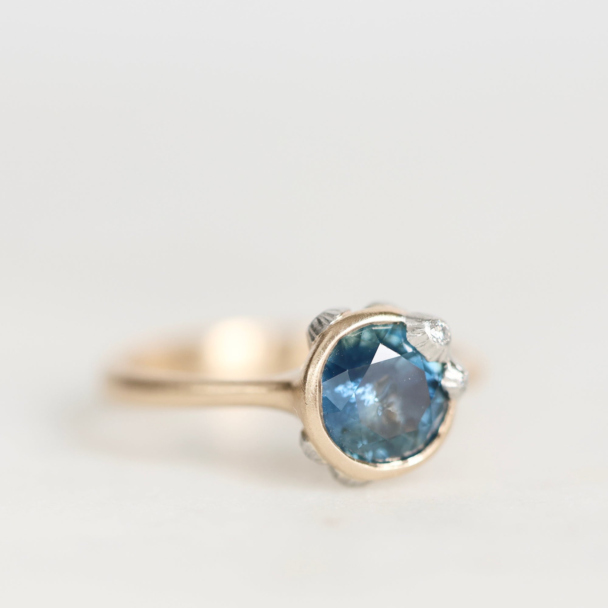 A close-up shot of a ring featuring a round-cut light blue gemstone in a silver-toned setting with small accent stones, on a plain light gray background.