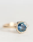 A close-up shot of a ring featuring a round-cut light blue gemstone in a silver-toned setting with small accent stones, on a plain light gray background.