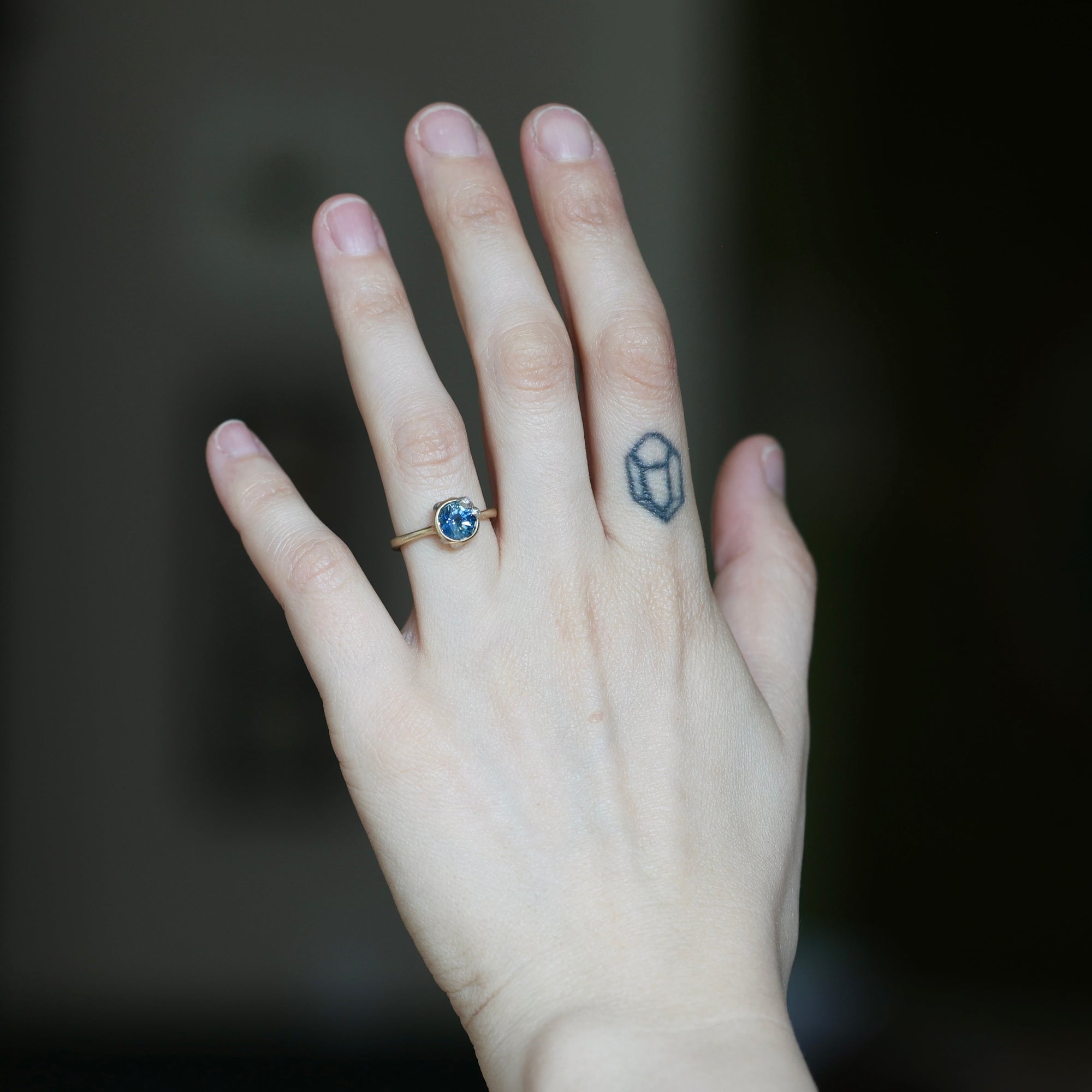 A close-up of a person's hand wearing a gold ring with a round blue gemstone, set against a dark, blurred background.