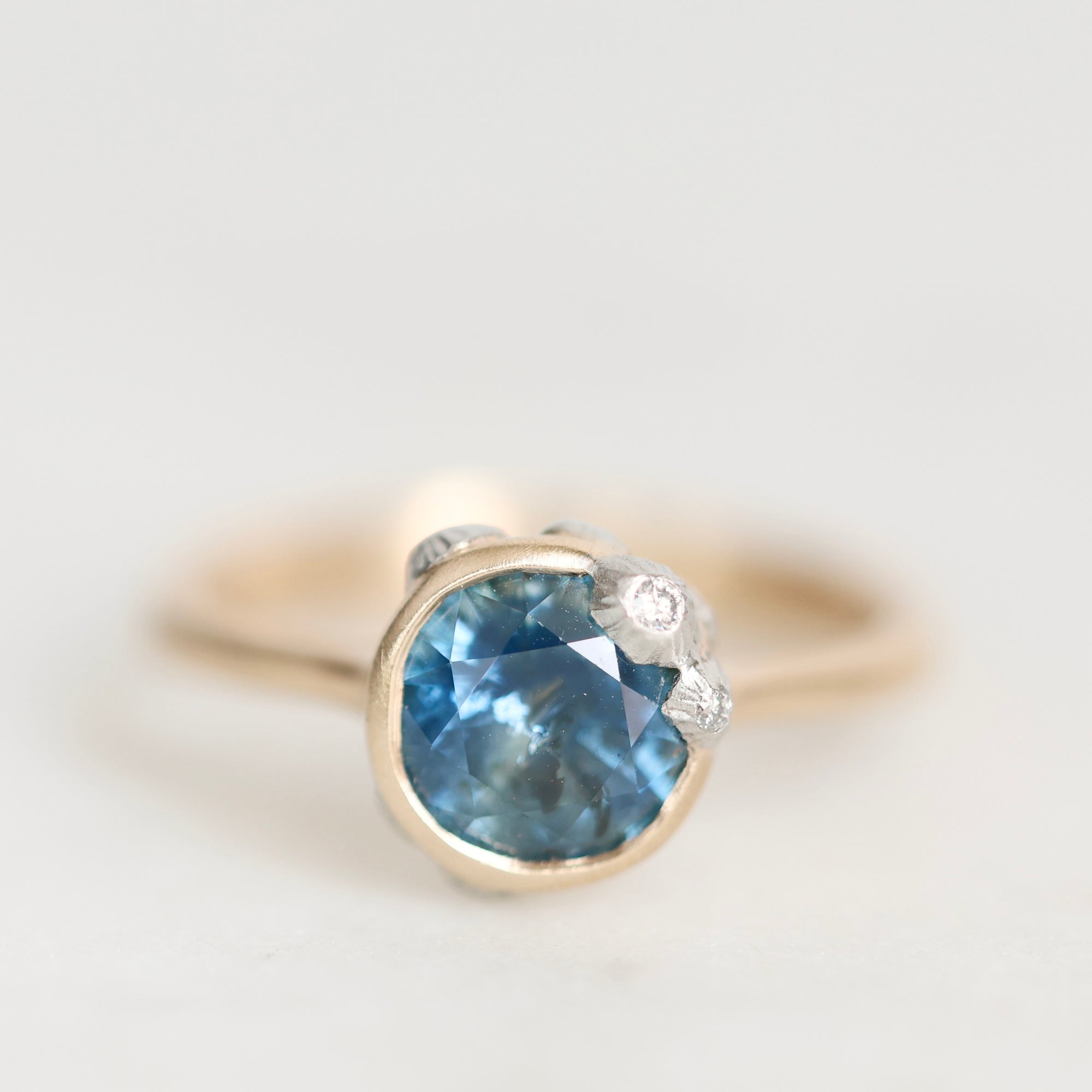 A close-up shot of a ring featuring a round-cut light blue gemstone in a gold bezel setting, accented by small clear stones on the shoulders, set against a plain light gray background.