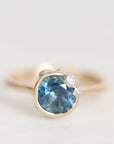 A close-up shot of a ring featuring a round-cut light blue gemstone in a gold bezel setting, accented by small clear stones on the shoulders, set against a plain light gray background.