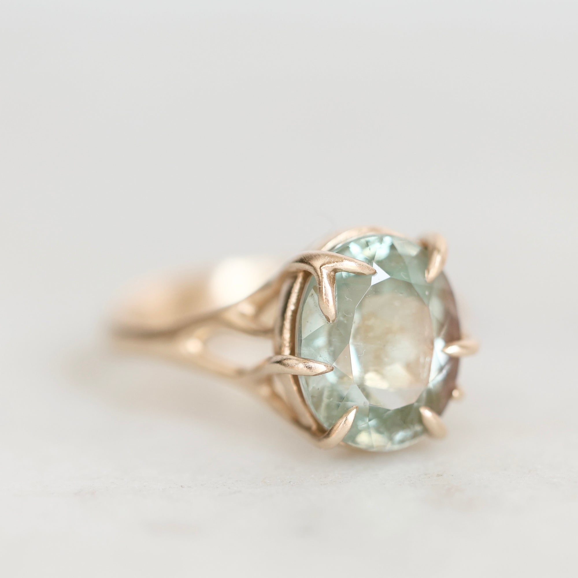 A close-up shot of a vintage-style ring featuring a large oval-cut light blue-green gemstone in a gold prong setting with leaf-like accents on the band, set against a soft, light gray background.