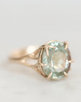 A close-up shot of a vintage-style ring featuring a large oval-cut light blue-green gemstone in a gold prong setting with leaf-like accents on the band, set against a soft, light gray background.