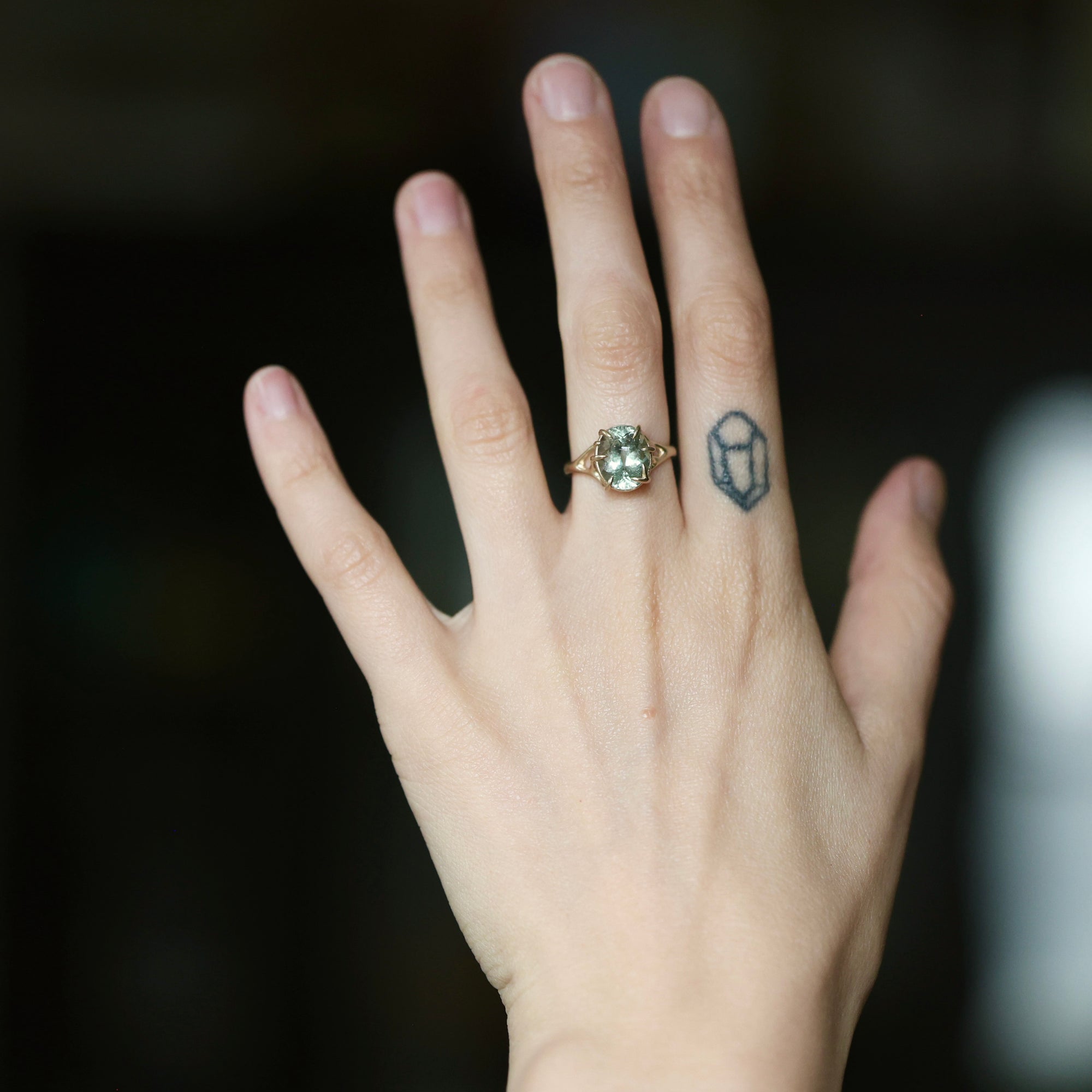 A close-up of a person's hand wearing a ring with a large oval light green gemstone in a gold setting, with a small geometric tattoo on the adjacent finger against a dark blurred background.