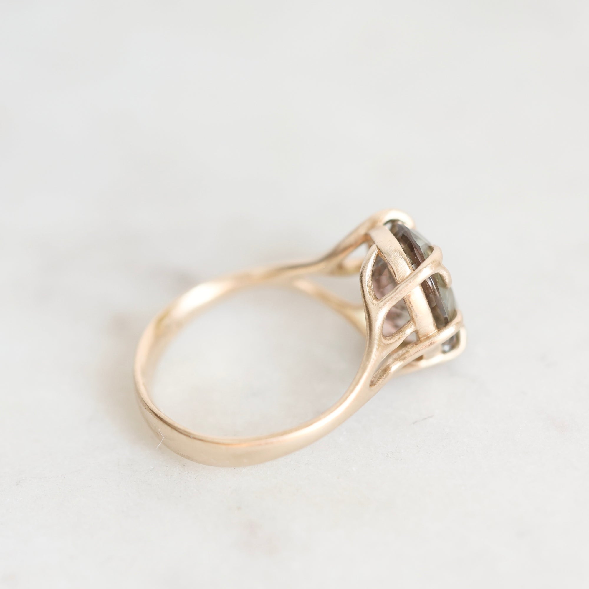A close-up shot of a gold ring with a basketweave setting holding a smoky, translucent gemstone, resting on a light marble surface.