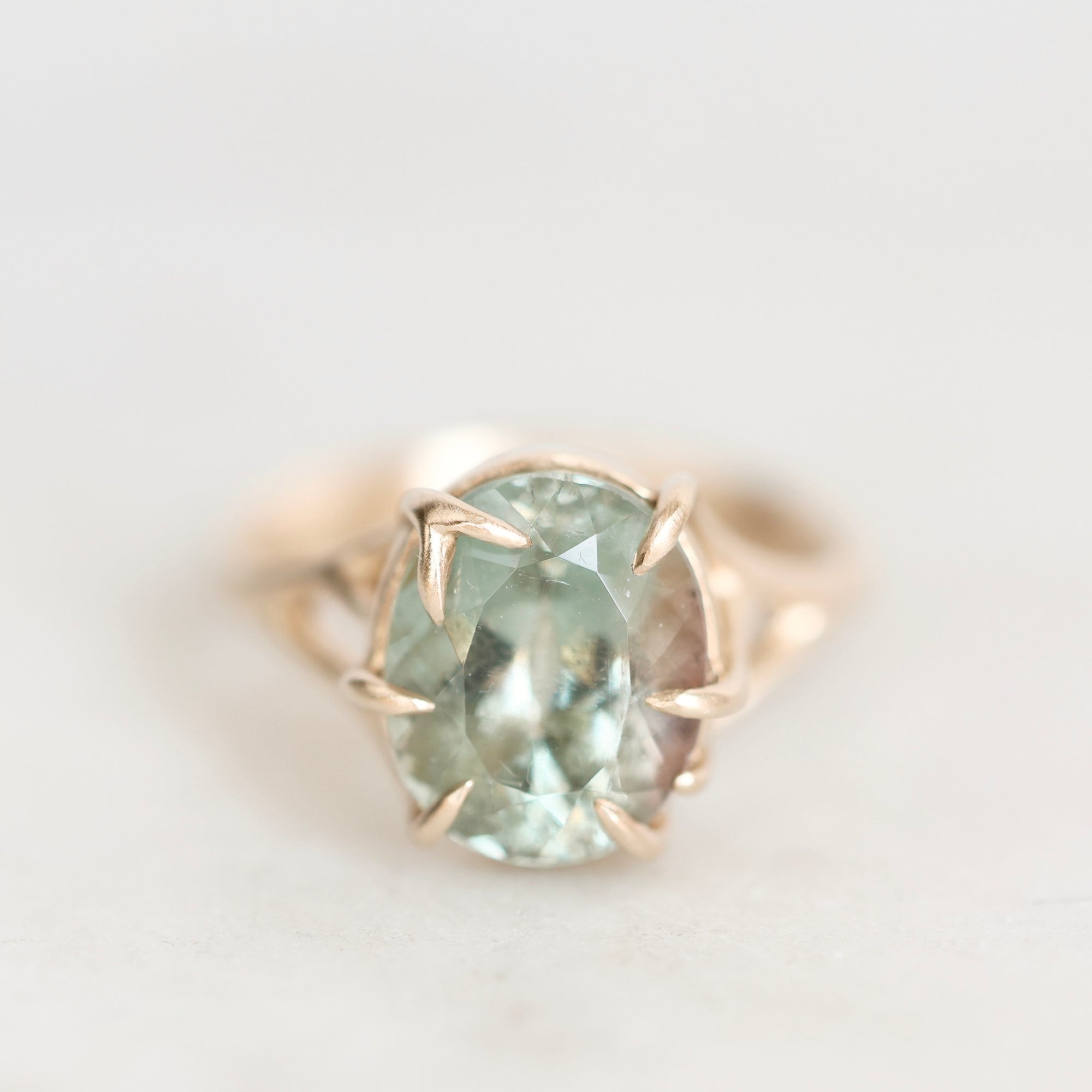 An oval-cut light green gemstone ring set in a rose gold prong setting, shown on a soft, light gray background.
