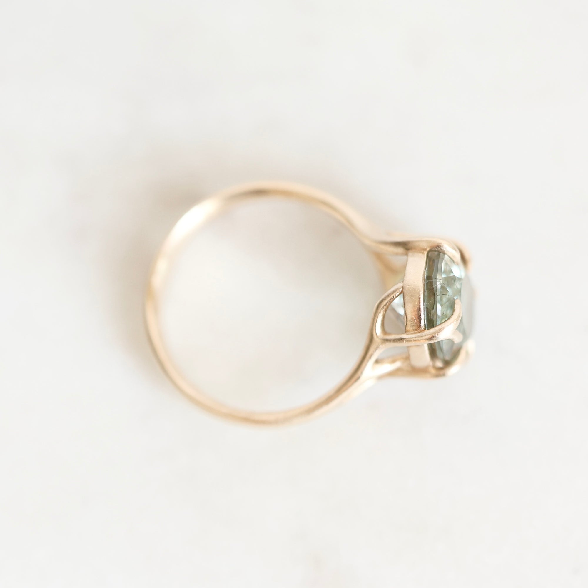 A gold ring with a twisted band and a bezel-set light green gemstone, photographed from above on a light off-white background.