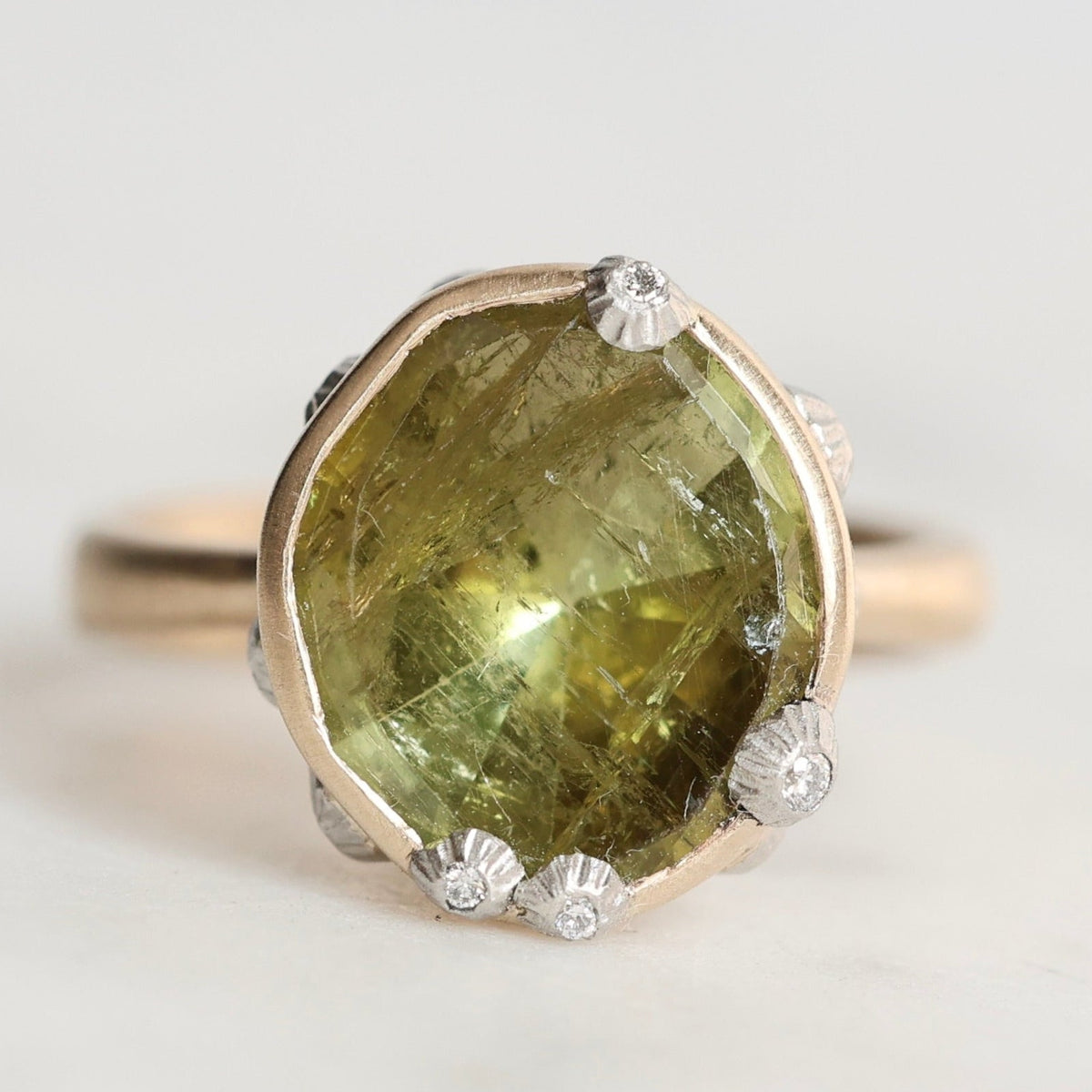 A gold ring featuring a large, irregular-cut olive green gemstone in a bezel setting, accented with small barnacles, photographed on a light gray background.