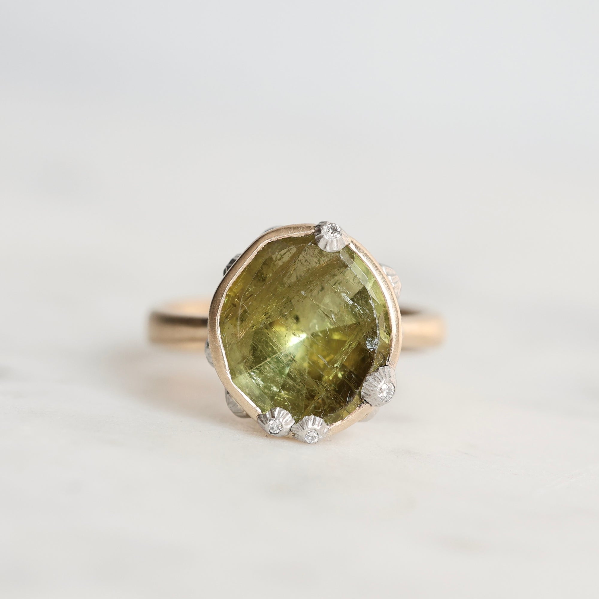 A gold ring featuring a large, irregular-cut olive green gemstone in a bezel setting, accented with small barnacles, photographed on a light gray background.