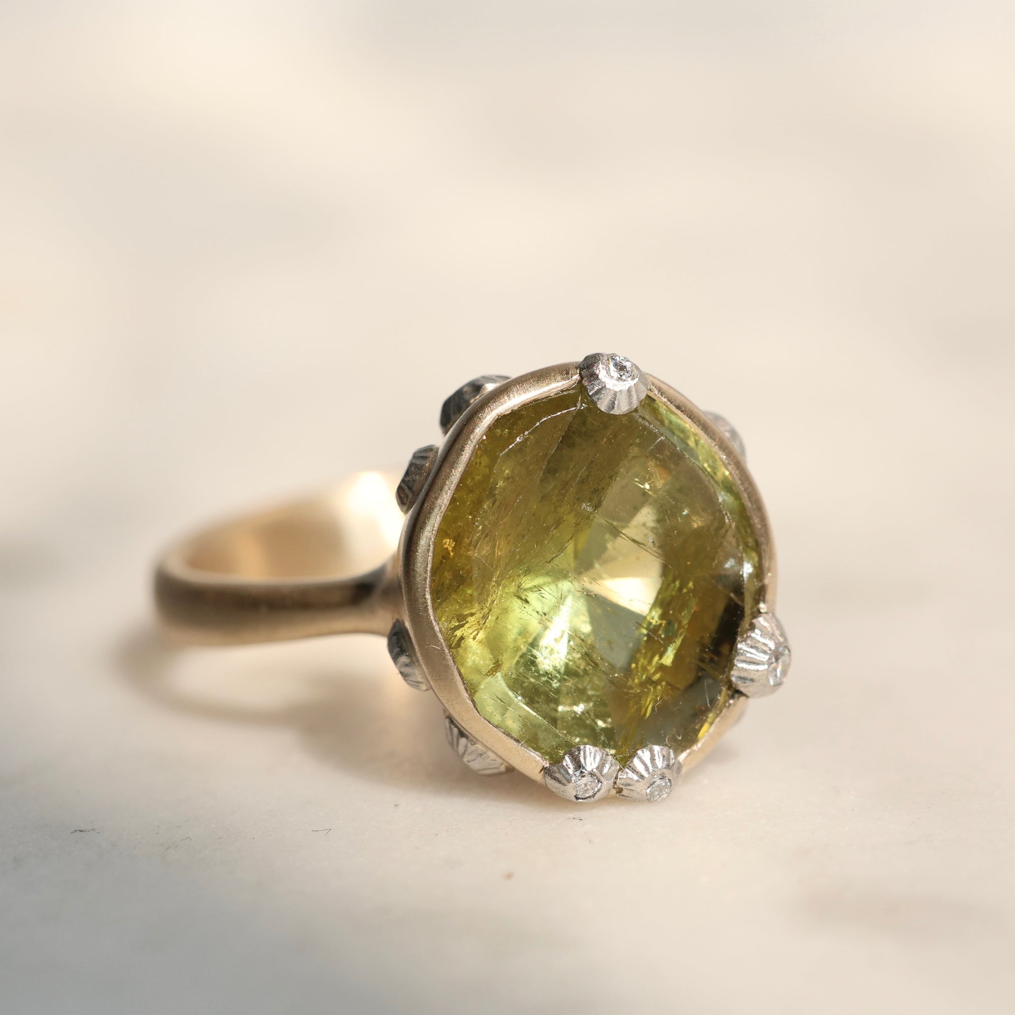 A silver ring featuring a large, faceted olive-green gemstone in a prong setting, with small barnacles along the band, photographed on a light beige background.