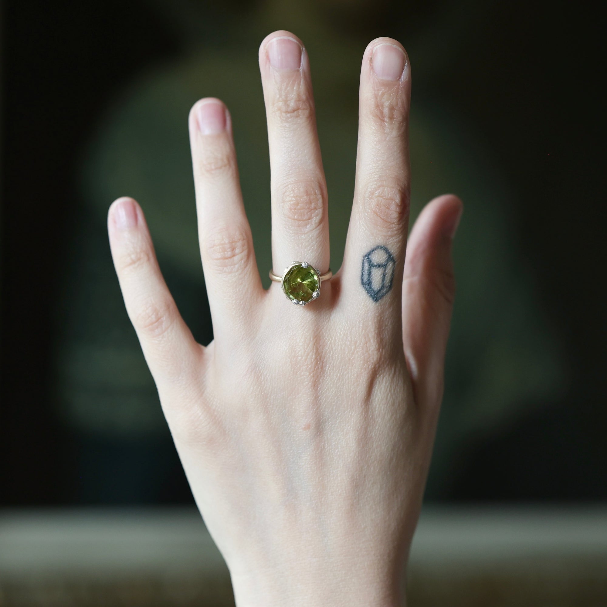 A hand wearing a gold ring with a round green gemstone, shown against a dark blurred background. The hand also has a small geometric tattoo on the middle finger.