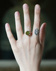 A hand wearing a gold ring with a round green gemstone, shown against a dark blurred background. The hand also has a small geometric tattoo on the middle finger.