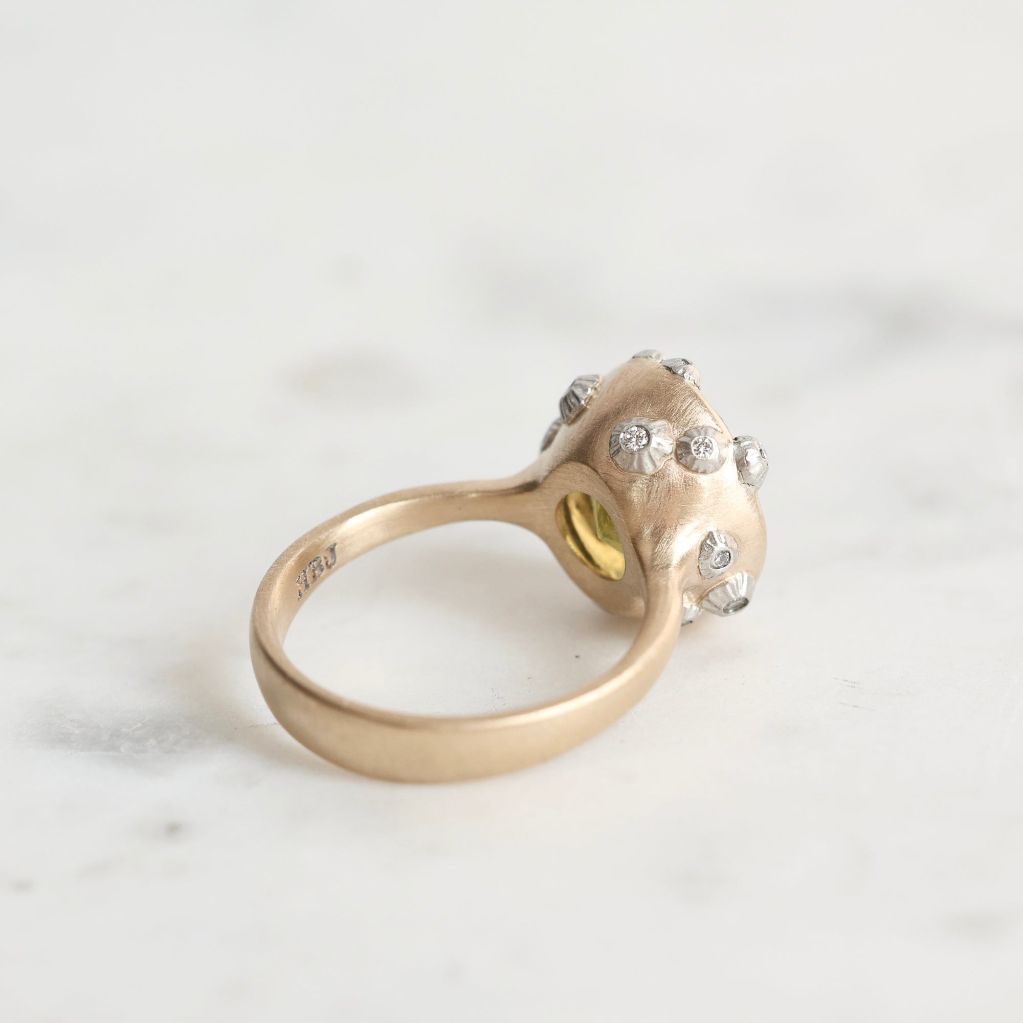 A gold ring with a textured, organic-shaped setting adorned with small barnacles, photographed on a light marble surface.