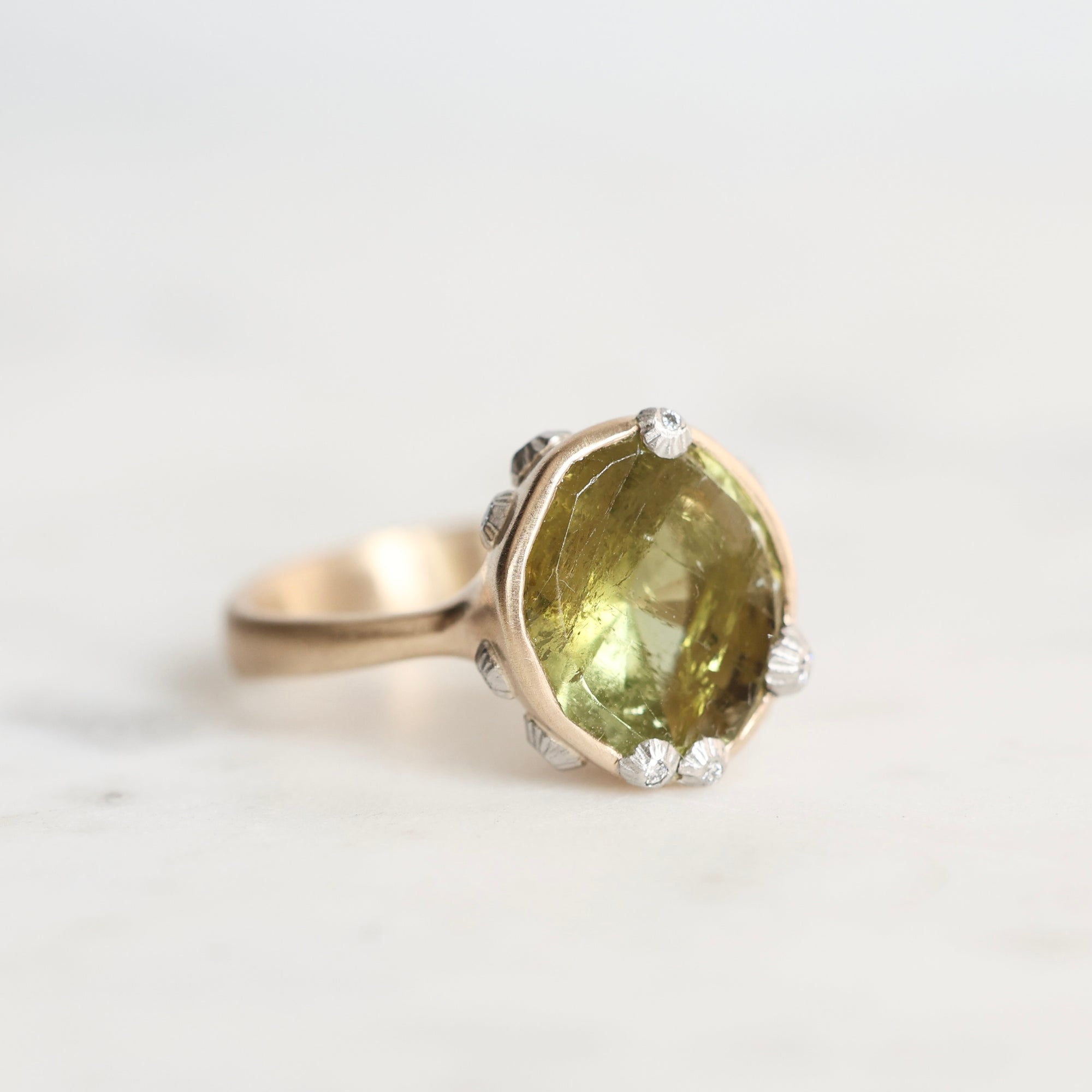 A vintage-style ring featuring a large, irregular-cut olive green gemstone in a gold-toned setting, accented with barnacles around the perimeter, photographed on a light marble surface.