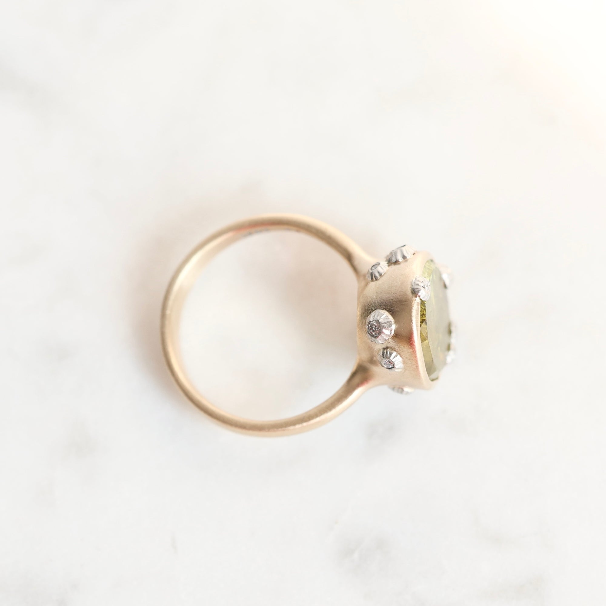 A gold ring with a round bezel-set pale green stone and small clear accent stones, photographed on a light marble surface.