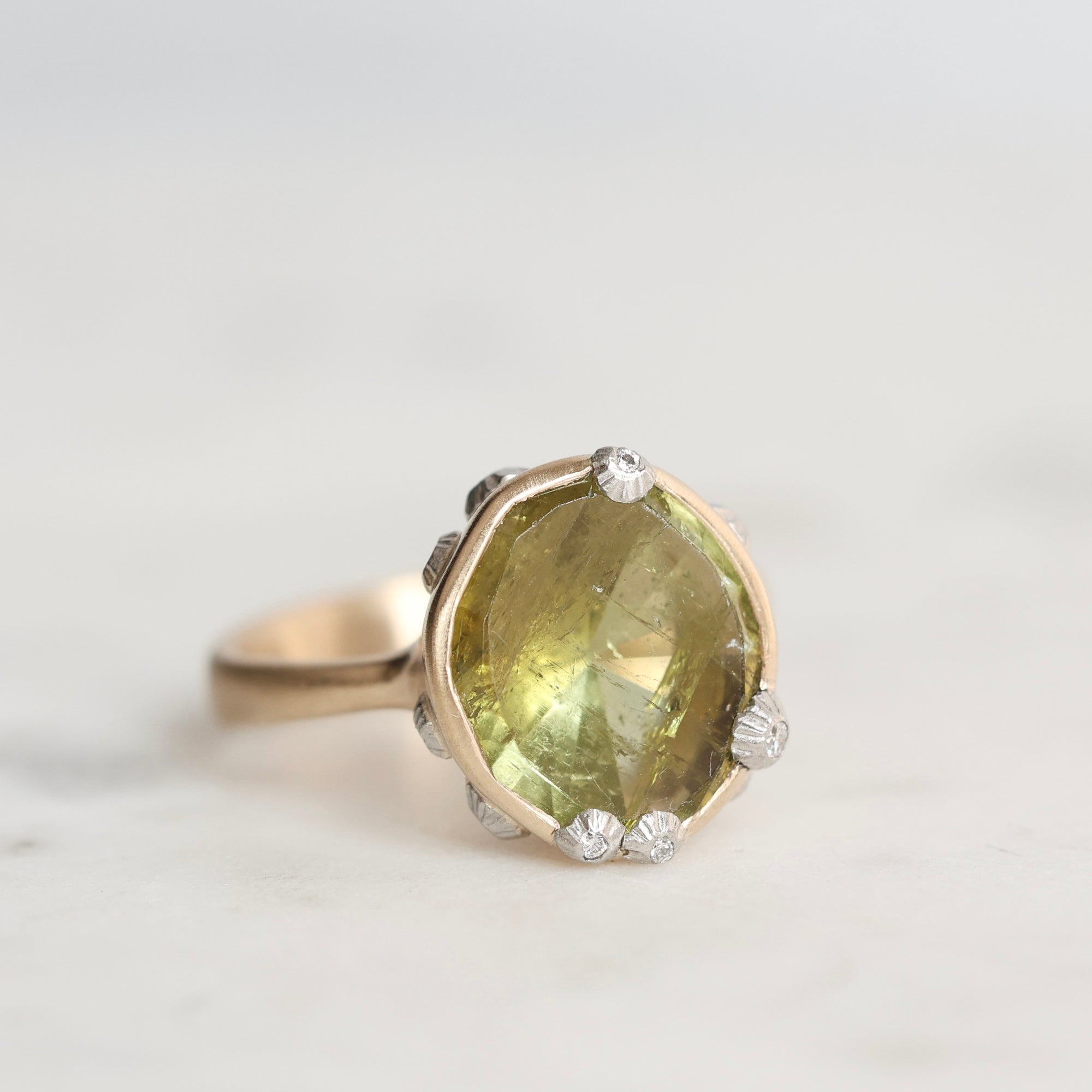 A gold ring featuring a large, faceted olive-green gemstone surrounded by small clear accent stones, set against a soft, light gray background.