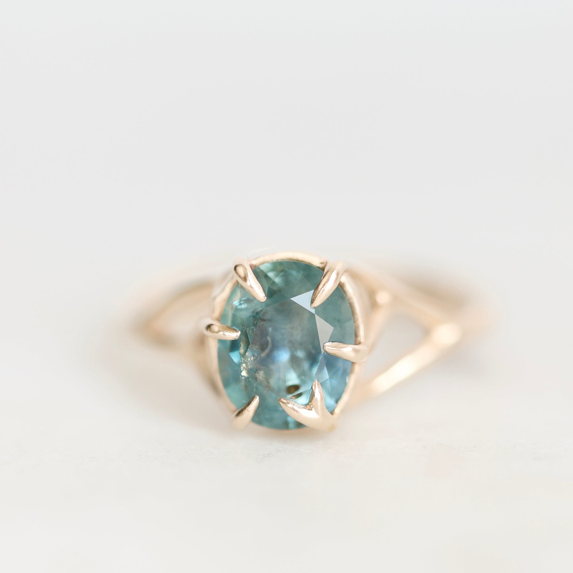 A gold ring featuring an oval-cut light blue gemstone in a prong setting, shown against a soft, light gray background.
