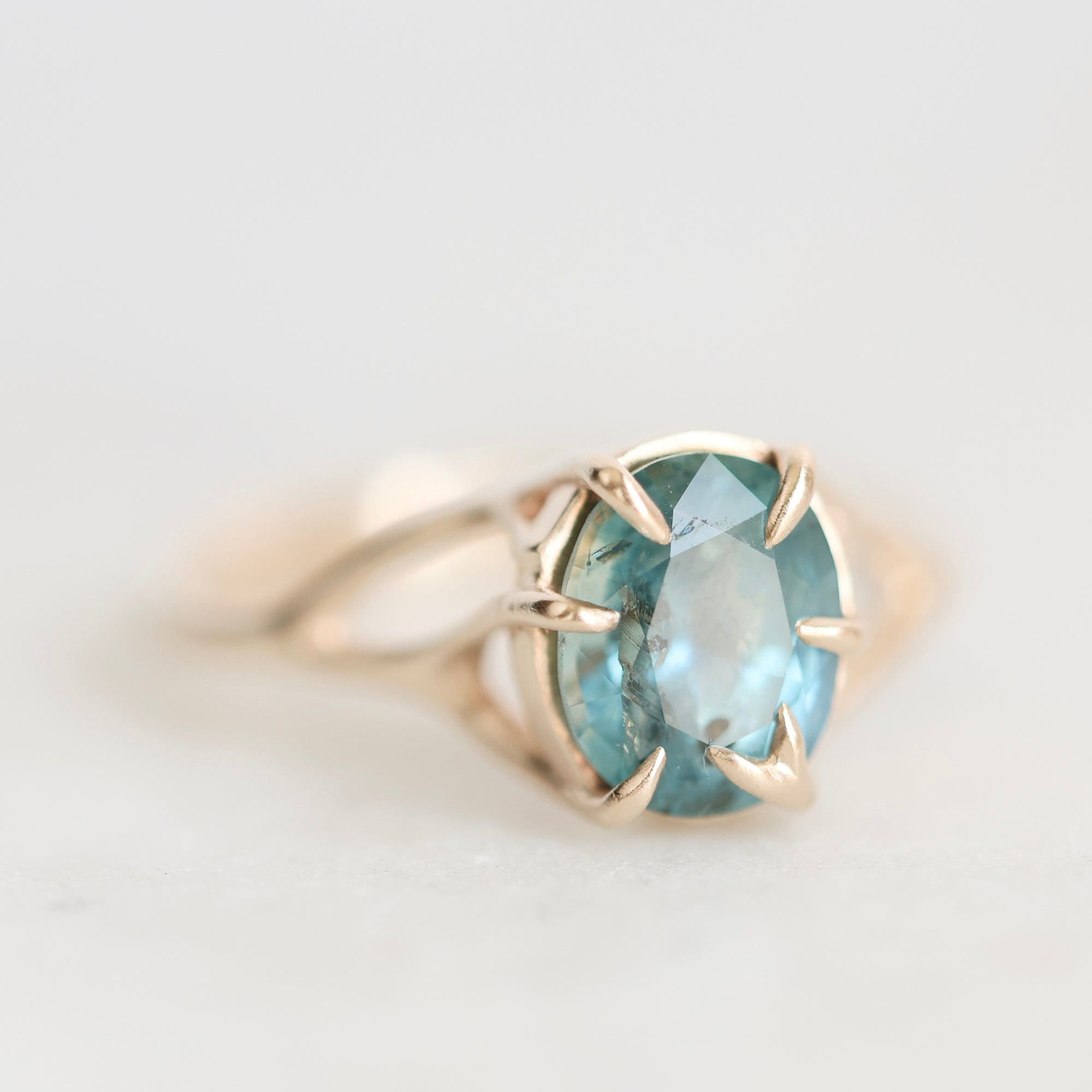 An oval-cut light blue gemstone ring set in a gold prong setting with a gold band, shown on a light gray background.