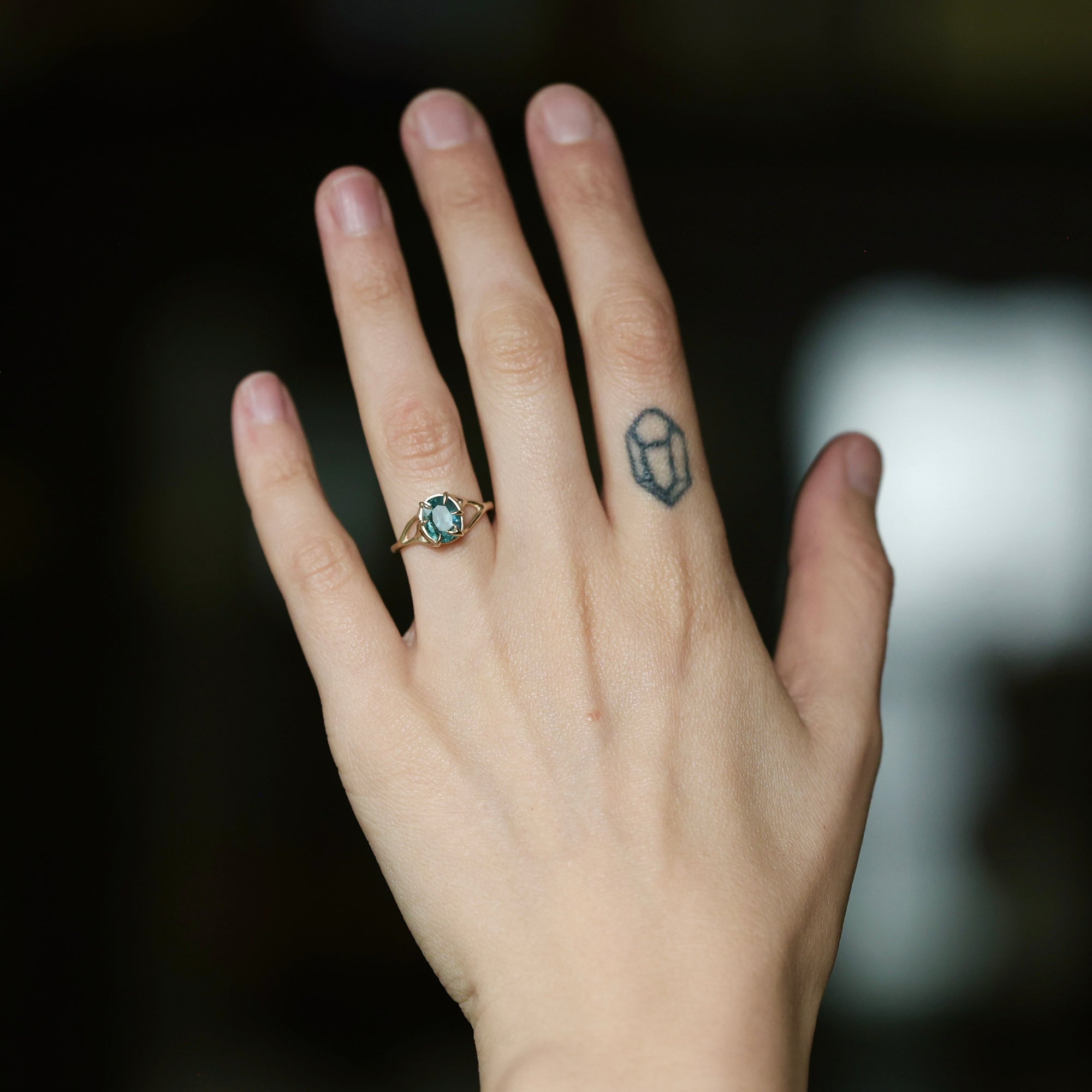 A close-up of a person's hand wearing a gold ring with a round turquoise-blue gemstone, set against a dark, blurred background.