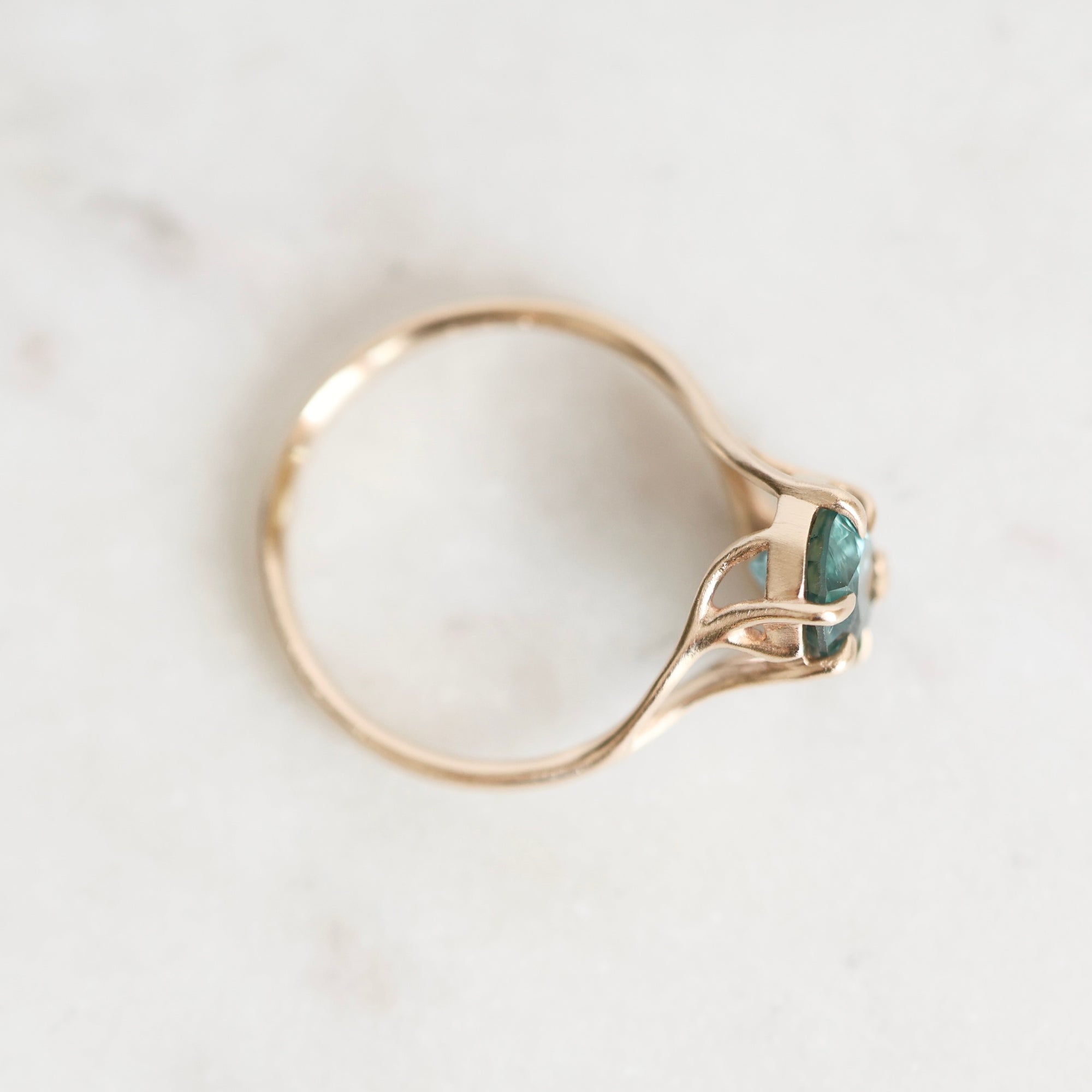 A delicate gold ring with a curved, openwork band and a small oval green gemstone, resting on a light marble surface.
