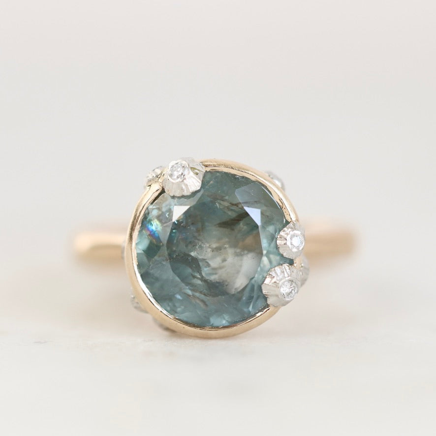 A round-cut light blue gemstone ring in a gold setting with small clear accent stones, photographed on a clean white background.