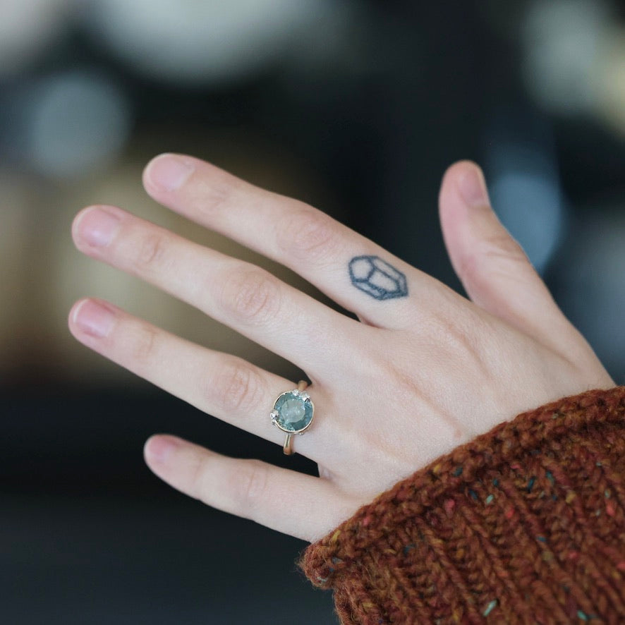 A close-up of a person's hand wearing a gold ring with a round light blue-green stone, set against a dark, blurred background. The person is wearing a brown knit sweater.
