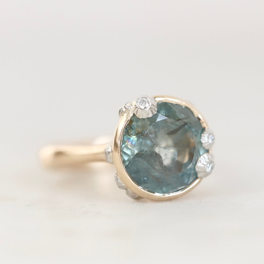 A close-up shot of a ring featuring a round, faceted light blue gemstone in a gold-toned setting, accented by small clear stones along the top and sides, on a light gray background.
