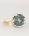 A close-up shot of a ring featuring a round, faceted light blue gemstone in a gold-toned setting, accented by small clear stones along the top and sides, on a light gray background.