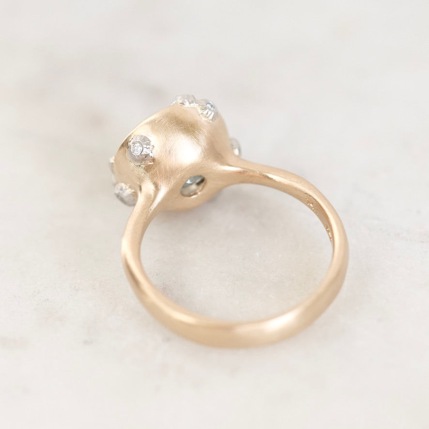 A close-up shot of a gold ring with a unique textured dome setting featuring small clear gemstones, resting on a light marble surface.