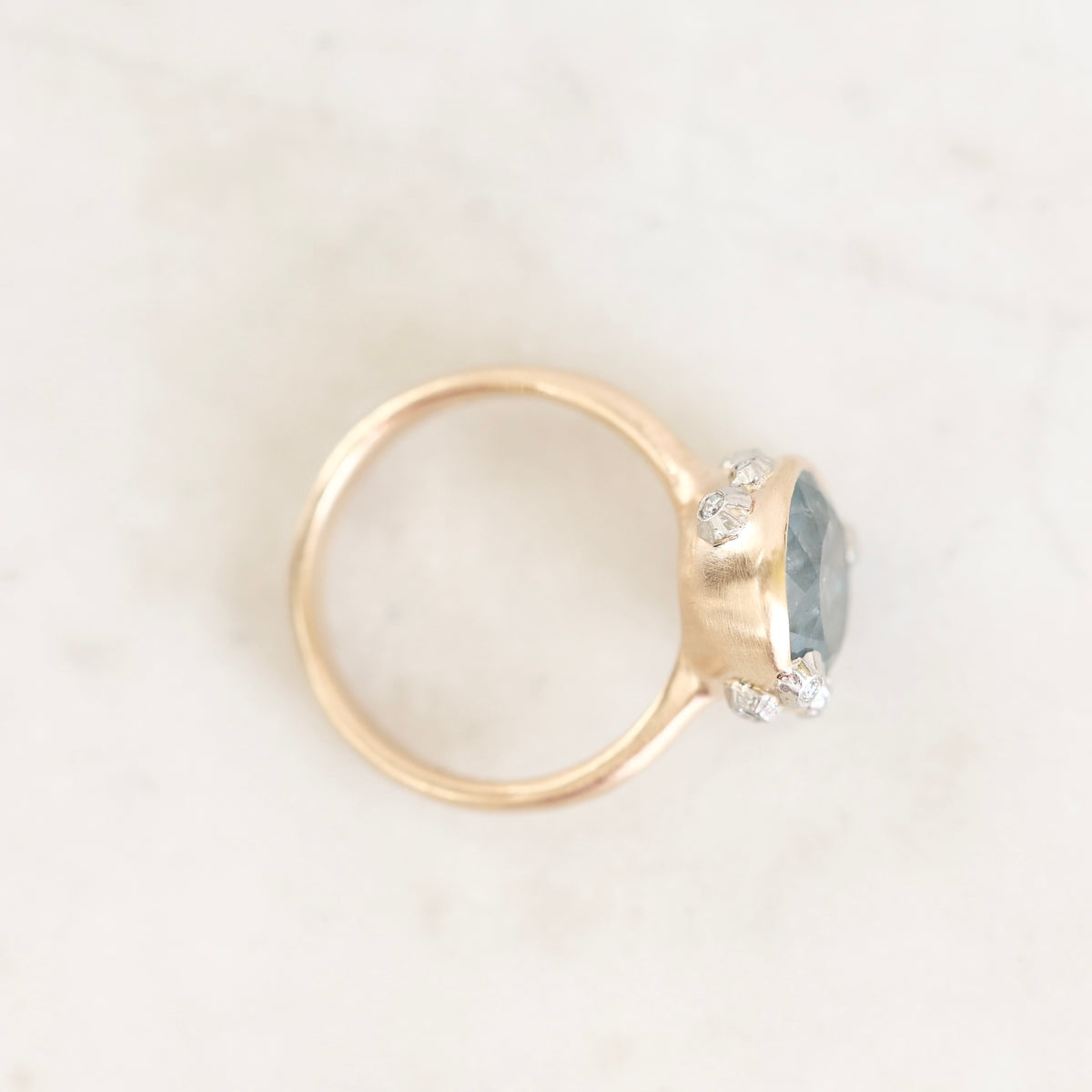 A gold ring with a round bezel holding a light blue-gray gemstone and small clear accent stones, photographed on a light marble-like surface.