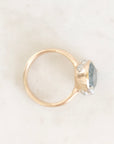 A gold ring with a round bezel holding a light blue-gray gemstone and small clear accent stones, photographed on a light marble-like surface.