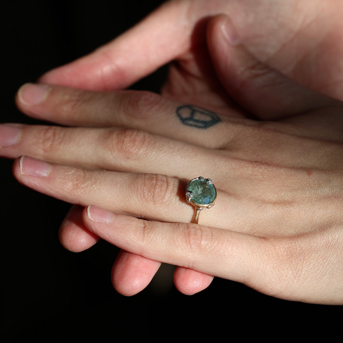 A close-up of a person's hand wearing a gold ring with a round, iridescent blue-green stone, set against a solid black background.