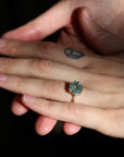 A close-up of a person's hand wearing a gold ring with a round, iridescent blue-green stone, set against a solid black background.