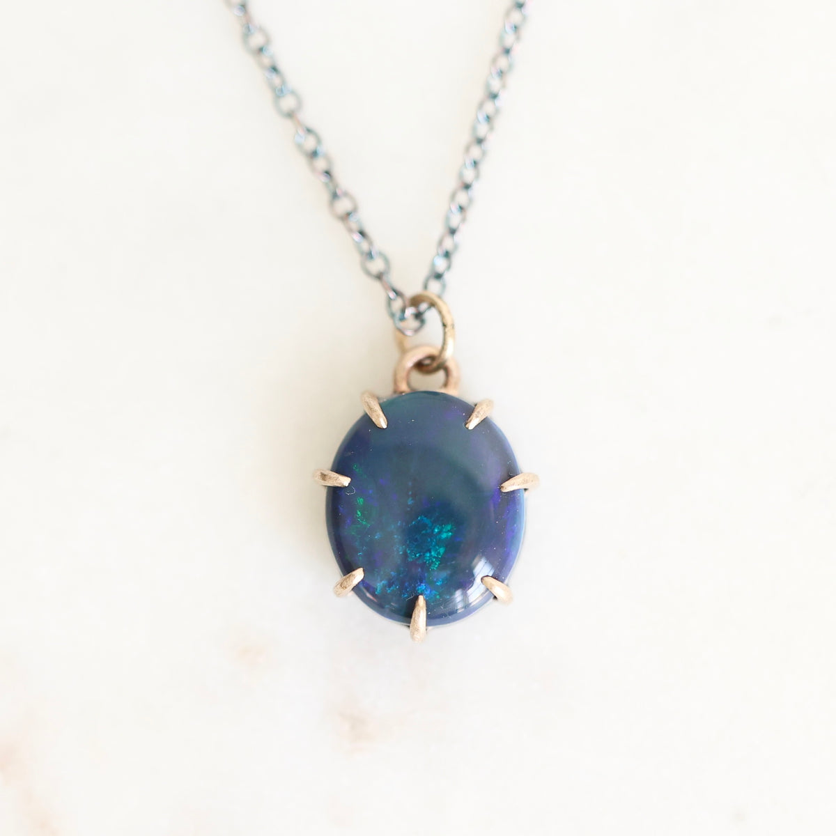Dark colored opal set in gold with an oxidized chain against a plain background. 
