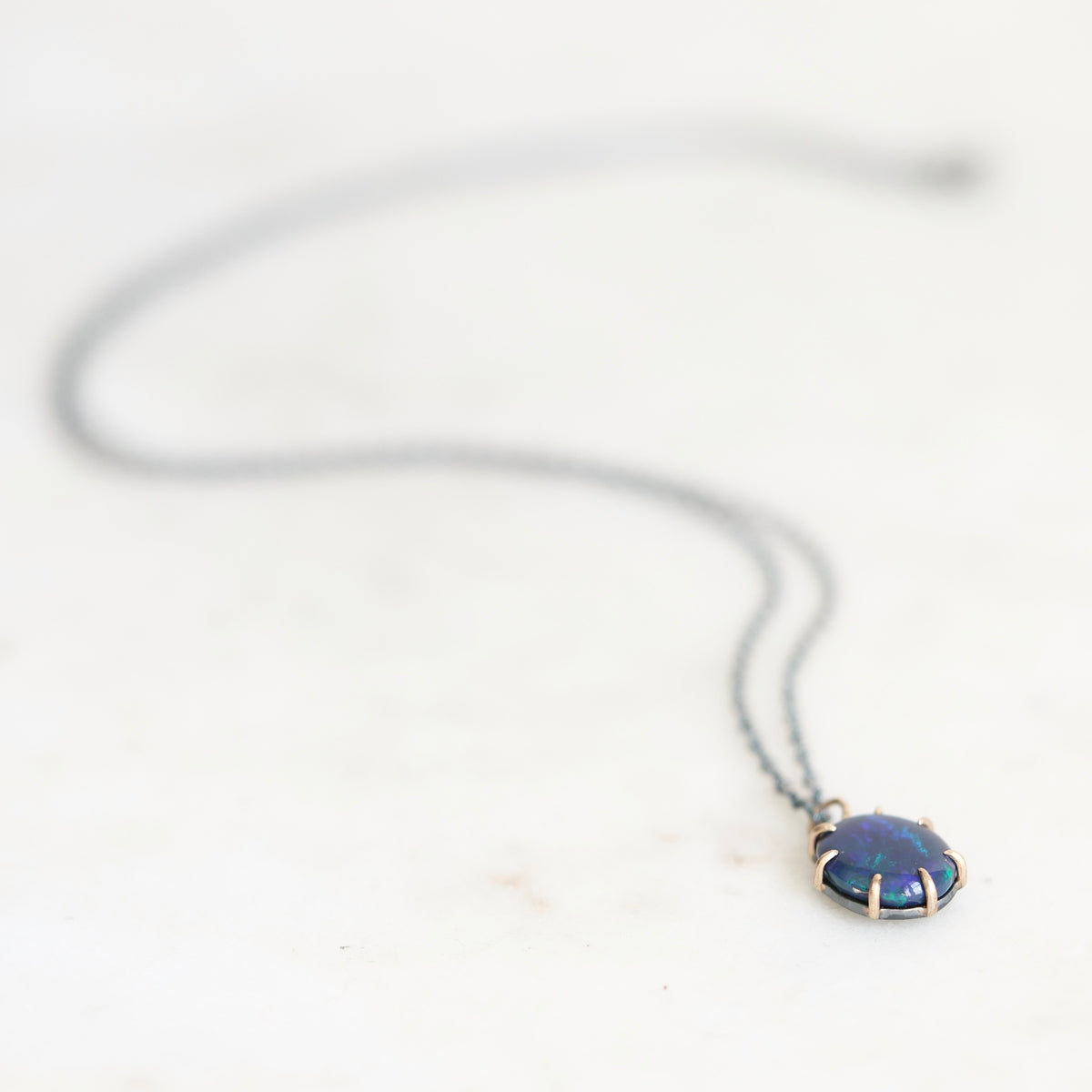 Dark colored opal set in gold prongs with an oxidized silver chain. Laying against a neutral background.