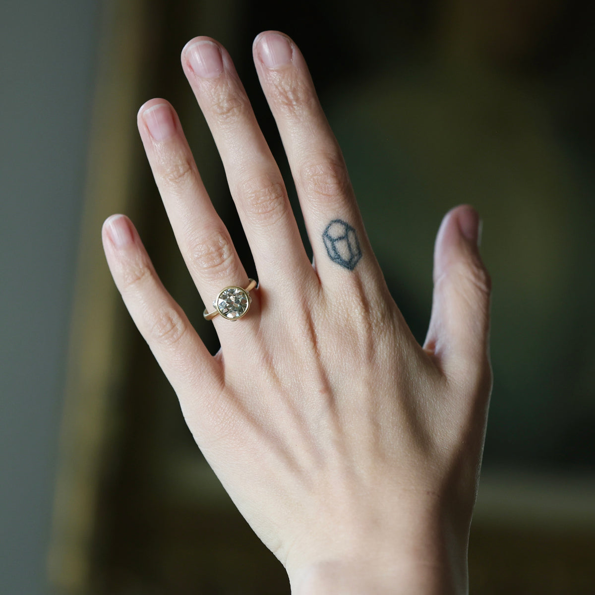 A close-up of a hand wearing a gold ring with a round clear gemstone, set against a softly blurred dark background.